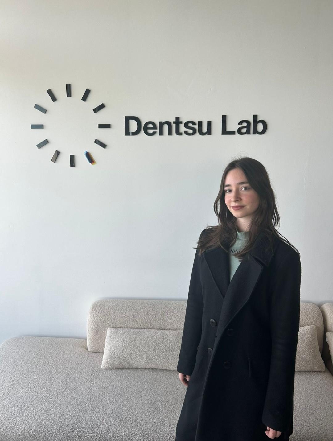 A person standing in front of a white wall with the Dentsu Lab logo, next to a beige sofa