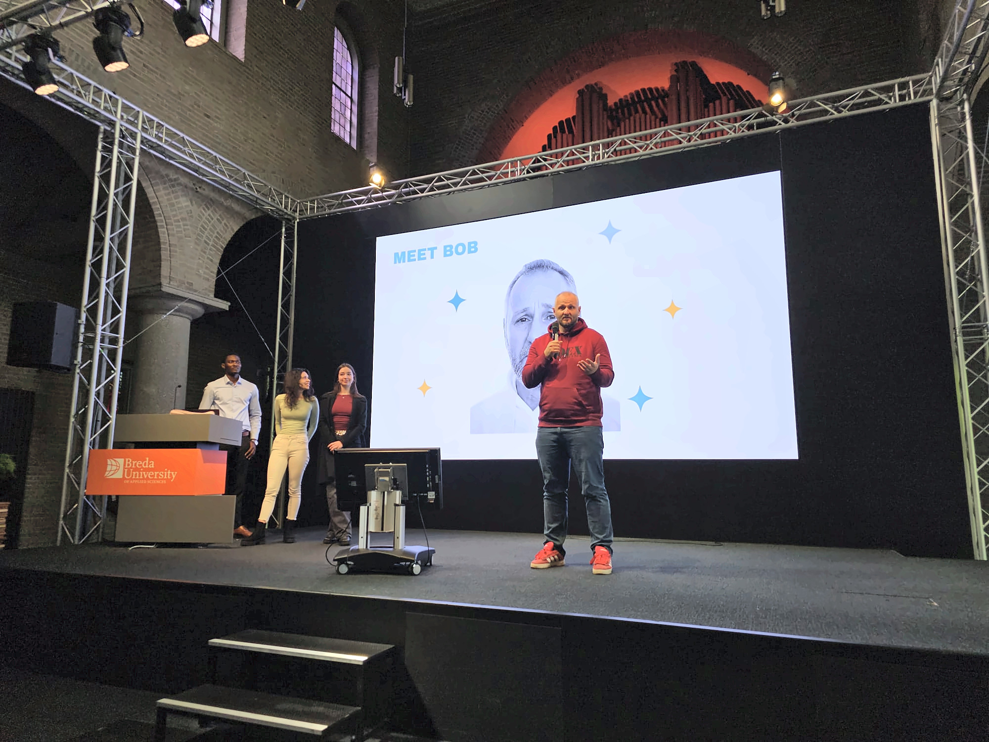 Three team members stand beside the lectern while a man speaks into a microphone in front of a large slide titled Meet Bob