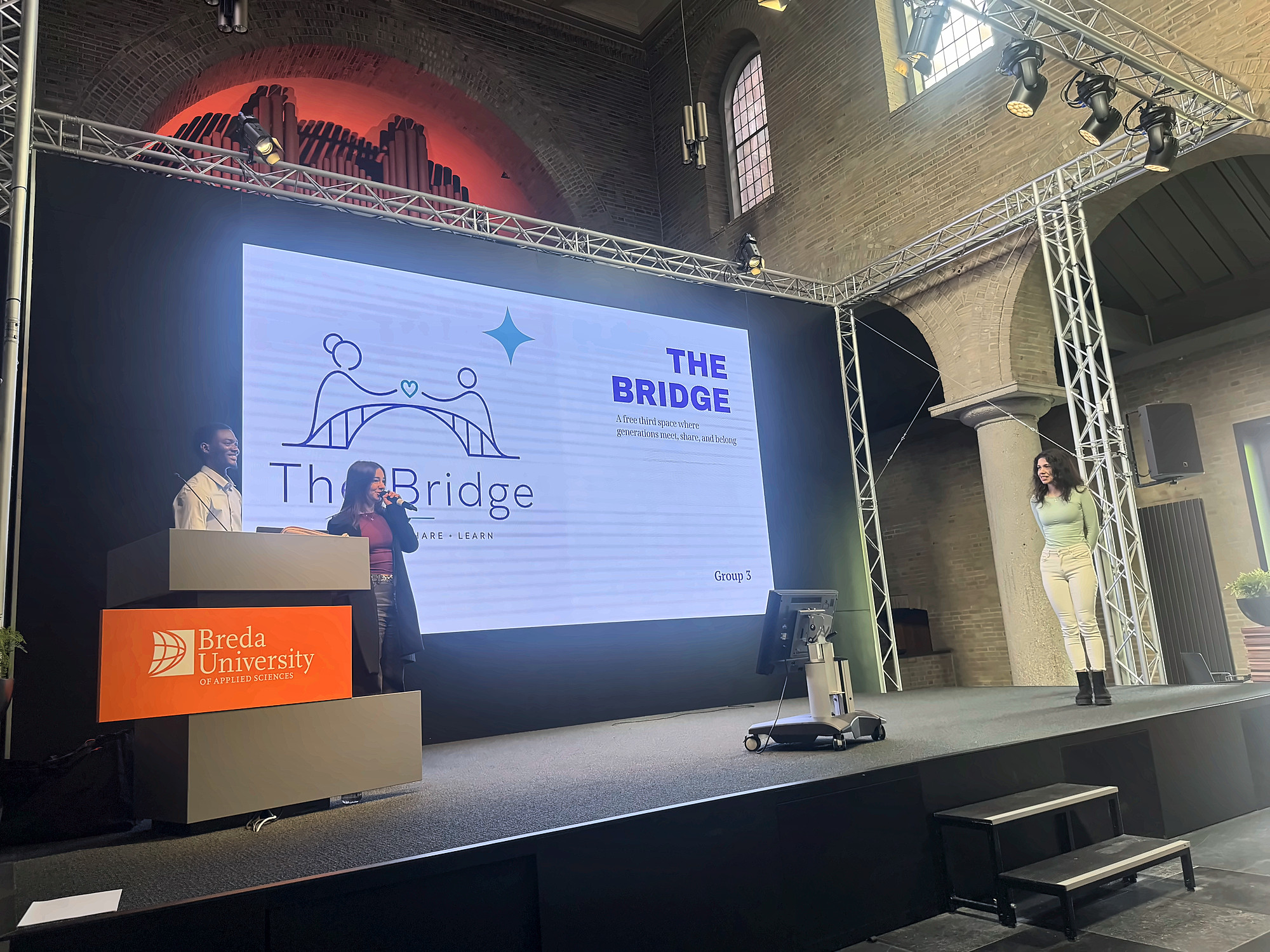 The BUas Chapel stage with the title slide of The Bridge on the screen, a presenter at the lectern and another standing beside the screen